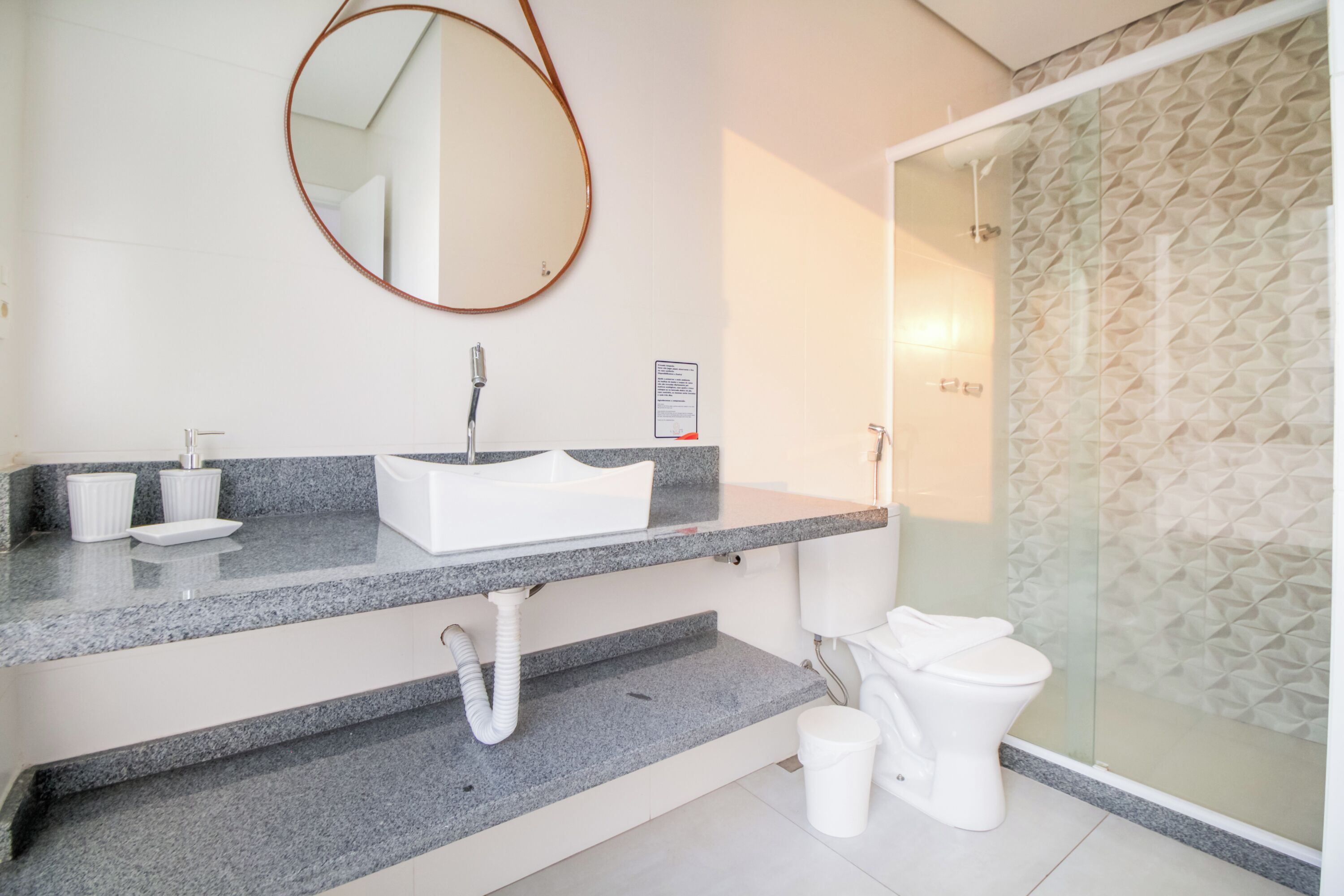 A modern bathroom with a granite countertop, a white sink, a round mirror, a toilet, and a shower with a patterned glass door.