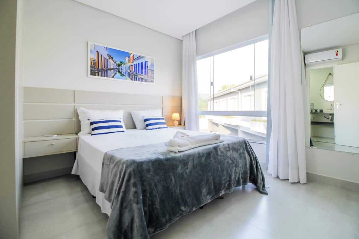 A hotel room with a bed, a nightstand, a window with curtains, and a wall-mounted air conditioning unit.