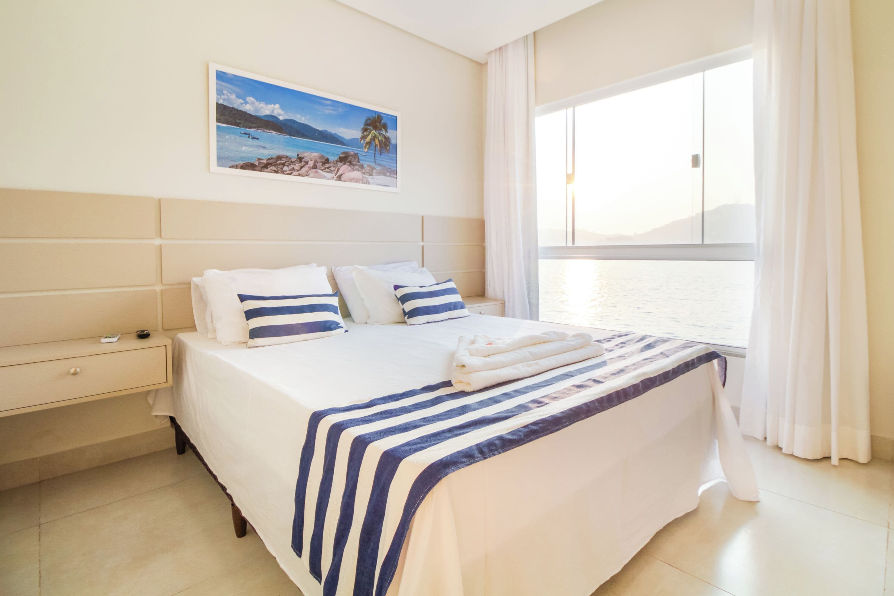 A hotel room with a large bed, a nightstand, a window with curtains, and a beach scene painting on the wall.