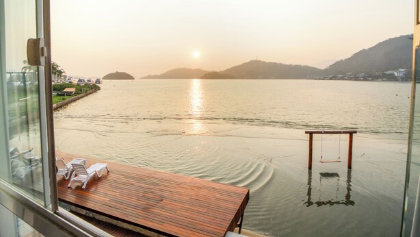 A wooden deck with lounge chairs and a swing, overlooking a calm body of water with a distant sun setting.