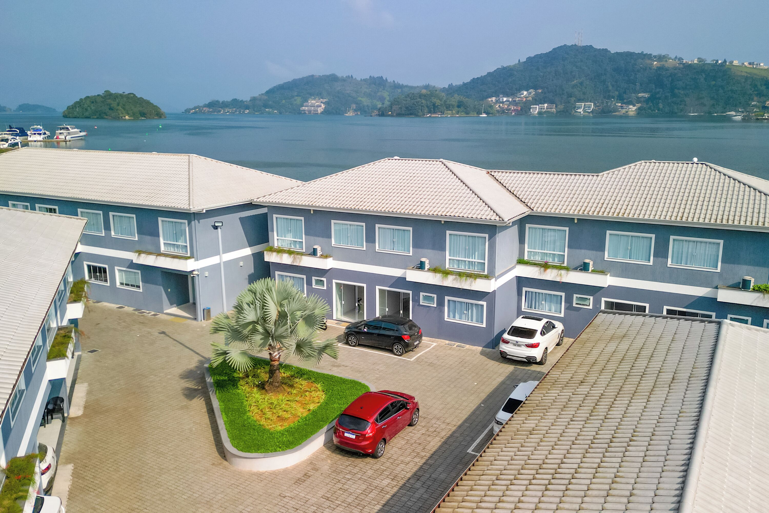 A modern building with a parking area, a palm tree, and a view of a lake and hills.