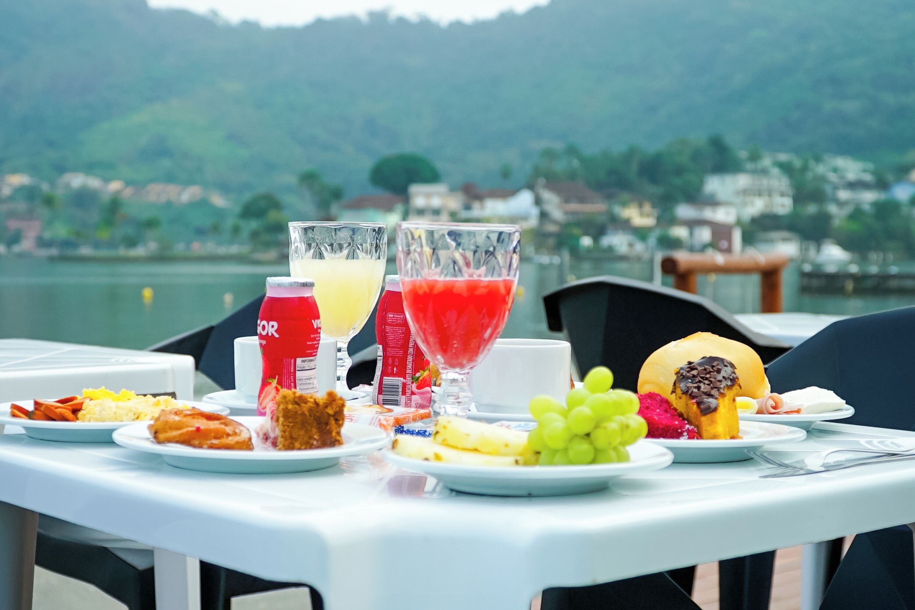 A table set with breakfast items including fruit, pastries, and beverages, overlooking a lake with buildings in the background.