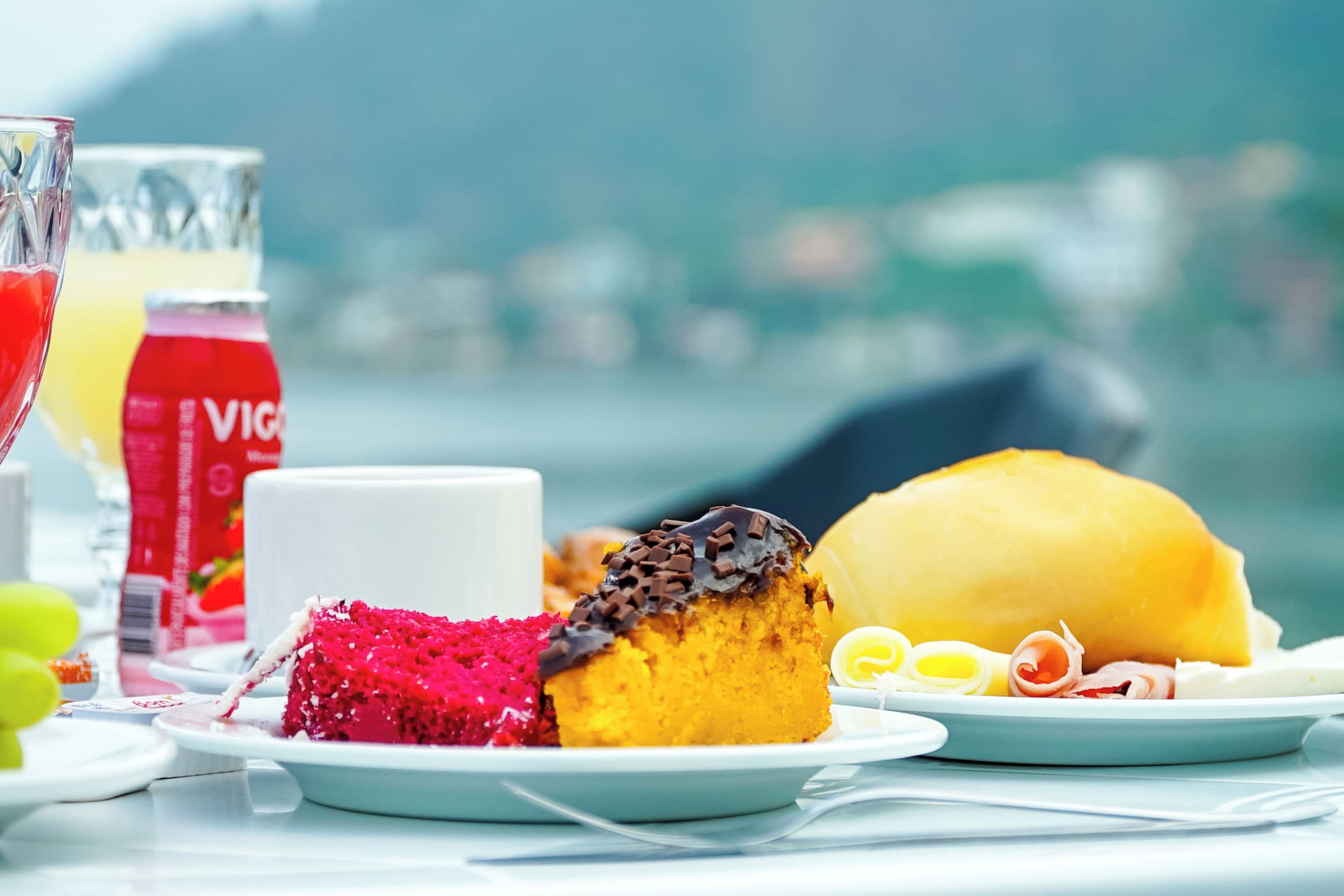 A table with a variety of food items including a slice of cake, a pastry, and some fruit, with a scenic view in the background.