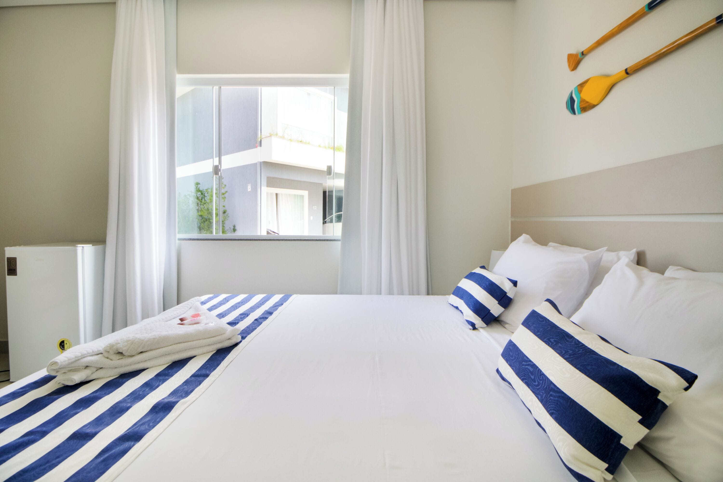 A hotel room with a large bed, white bedding, and striped pillows. There is a window with curtains, a view of buildings outside, and two wooden paddles on the wall.