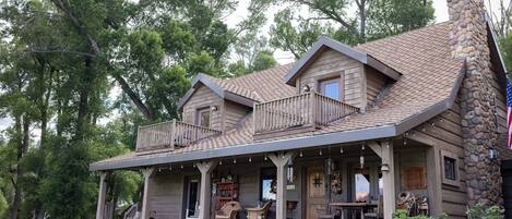 A two-story log cabin with a stone chimney, a porch with outdoor seating, and a view of the surrounding trees.