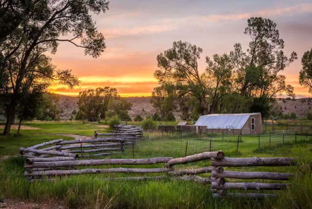 Une grange rustique avec un toit en métal, entourée d’une clôture en bois et d’arbres, se dressant sur fond de coucher de soleil.