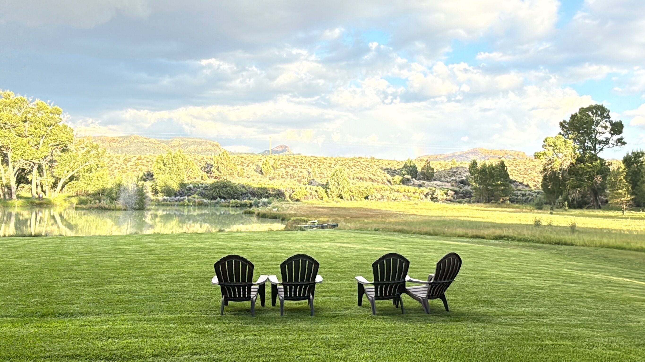 Un champ verdoyant avec trois chaises Adirondack disposées face à un lac, avec des collines en arrière-plan.