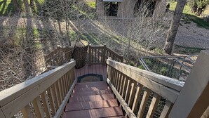Eine hölzerne Treppe führt zu einem Gebäude mit einem Metall-Dach, umgeben von Bäumen und einer grünen Wiese.