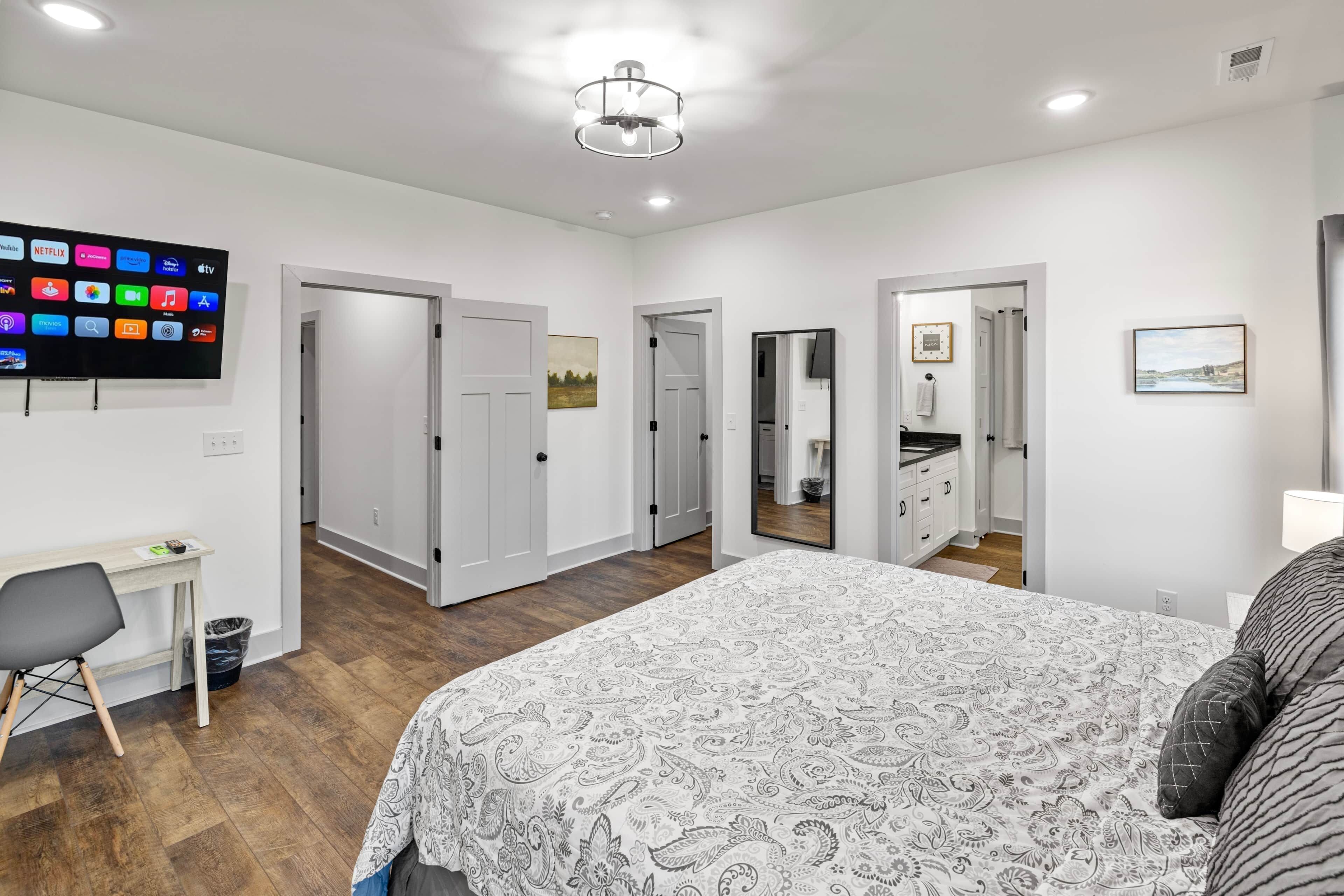 A hotel room with a bed, a desk, a chair, a flat-screen TV, and a bathroom visible through an open door.