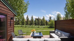 Eine Holzterrasse mit Gartenmöbeln, eine Glastür und Blick auf einen Rasen mit Bäumen und Bergen im Hintergrund.