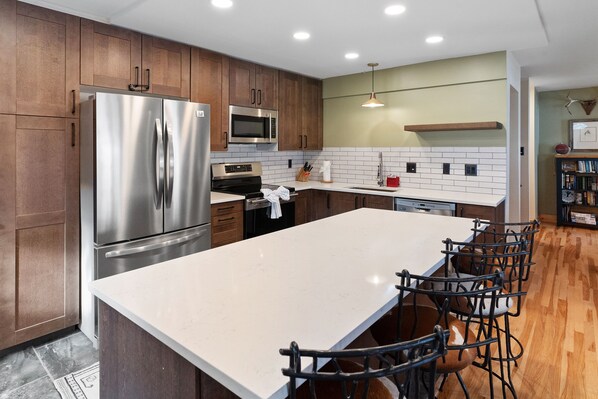 A modern kitchen with wooden cabinets, stainless steel appliances, a large island with bar stools, and a hanging light fixture.