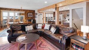 A cozy living room with a large sectional sofa, a coffee table, and a rug. There is a dining area with a table and chairs, and a kitchen in the background.