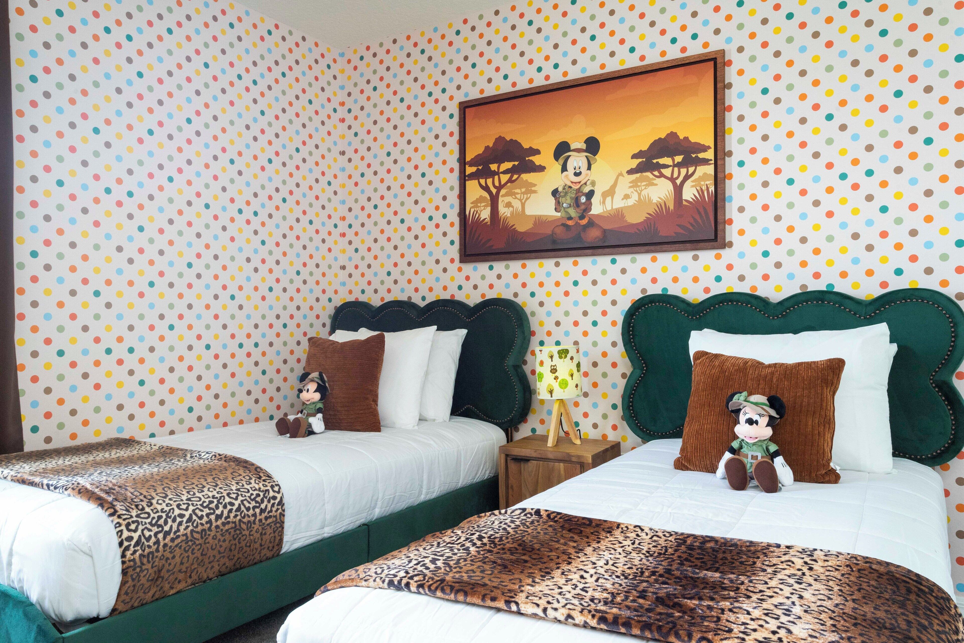 A hotel room with two beds, a polka dot wallpaper, a framed picture of Mickey Mouse, and a Mickey Mouse plush toy on one of the beds.