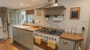 Een keuken met een fornuis, oven en een spoelbak. Er zijn keukenkastjes, een raam met gordijnen en een wandplank met spullen.