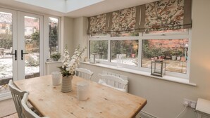 Een glazen veranda met een houten tafel, witte stoelen en een raambekleding met bloemenprint.