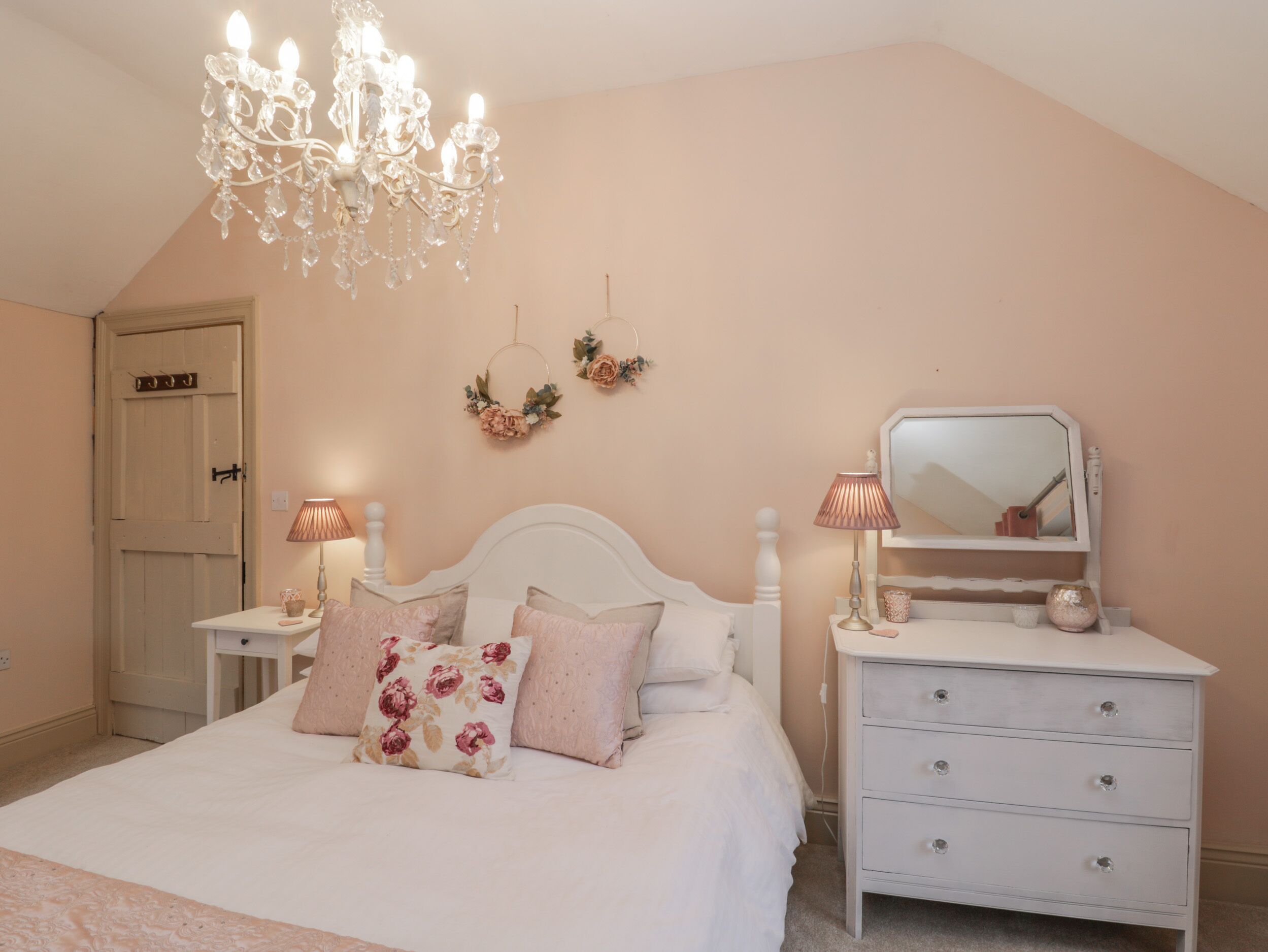 Une chambre à coucher avec un lit blanc, des coussins à motifs floraux, une commode blanche, un miroir et un lustre.