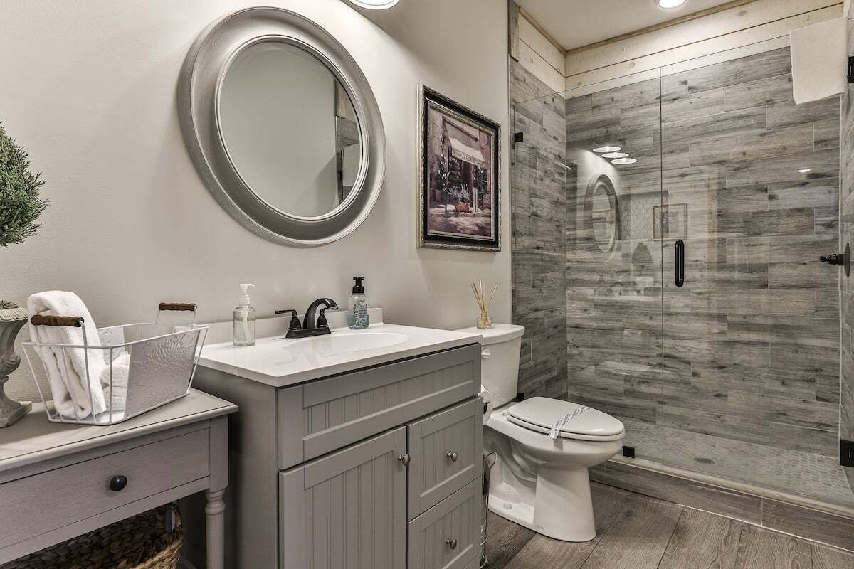 A bathroom with a round mirror, a toilet, a sink with a basket of towels, and a shower area with a glass door.
