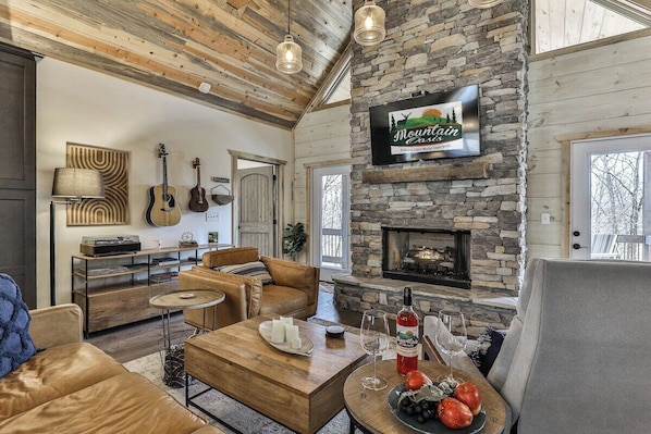 Un salon chaleureux avec une cheminée en pierre, des canapés en cuir et un téléviseur fixé au mur diffusant l’émission « Mountain Cabin ».