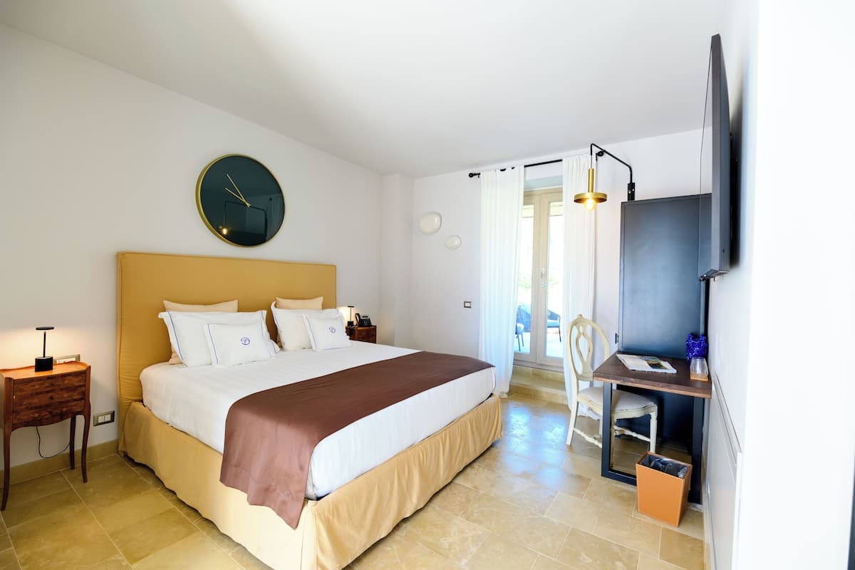 A bedroom with a bed, bedside table, clock, TV, and a desk with a chair.