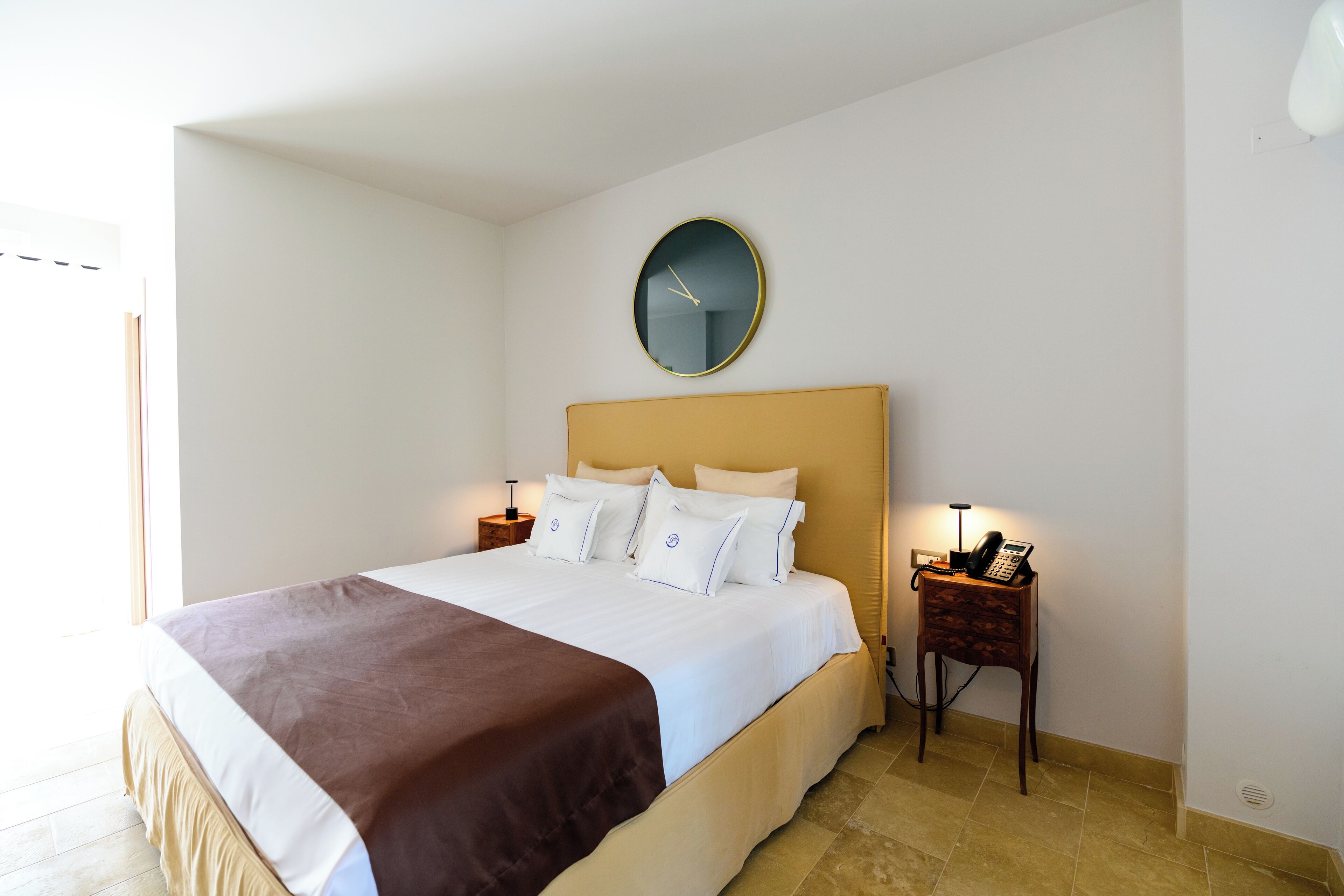 A neatly made bed with a brown throw blanket, a round wall clock, and a bedside table with a lamp and phone.
