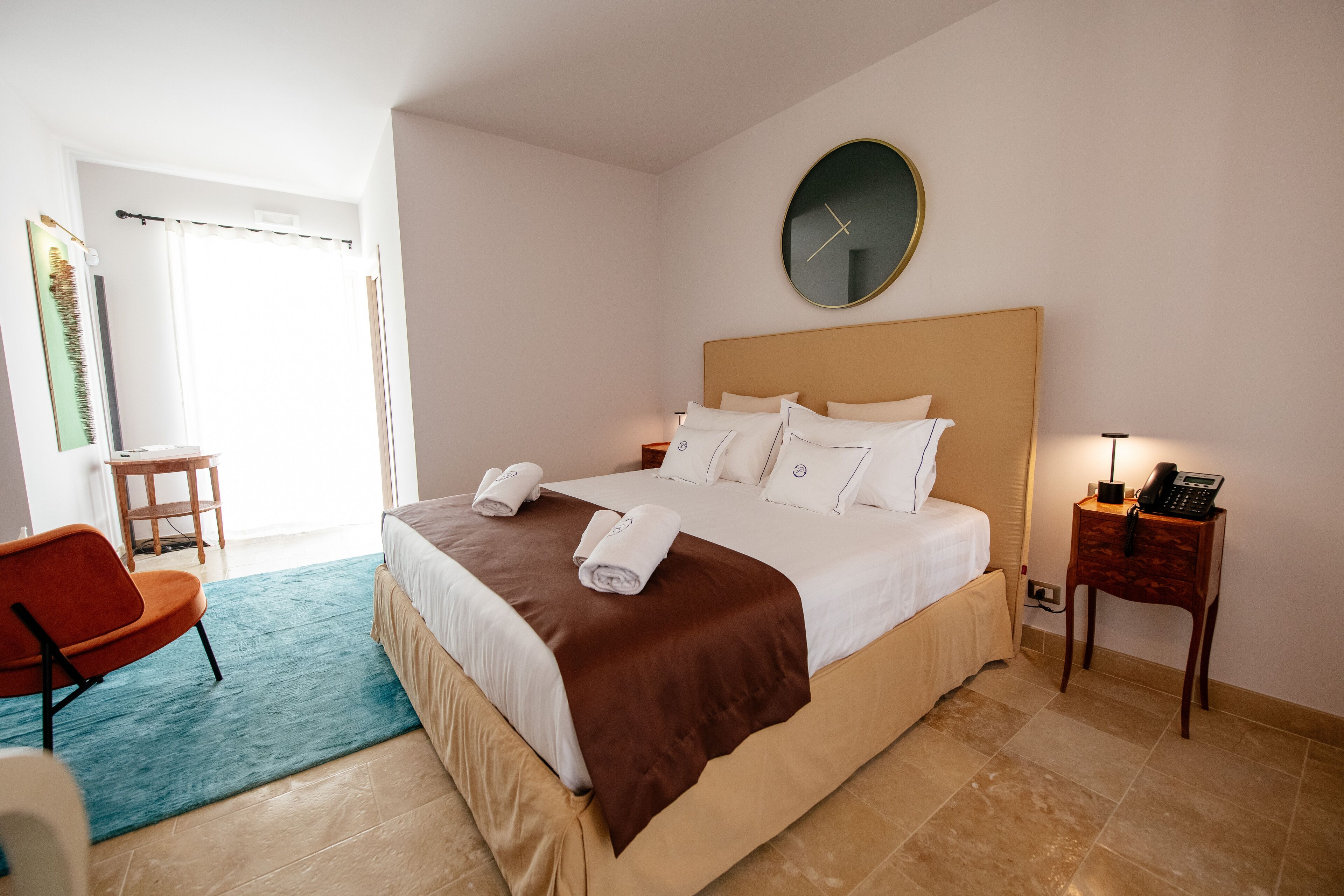 A hotel room with a bed, a chair, a bedside table with a lamp, and a clock on the wall.