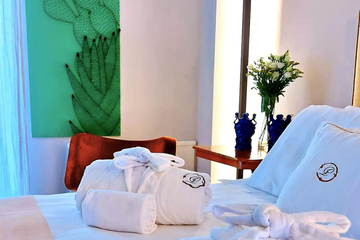 A hotel room with a bed, towels, a green wall art, a vase with flowers, and a wooden chair.