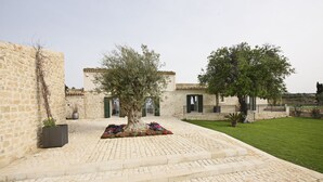 A stone building with a paved courtyard, steps leading to a door, and a grassy area in the background.