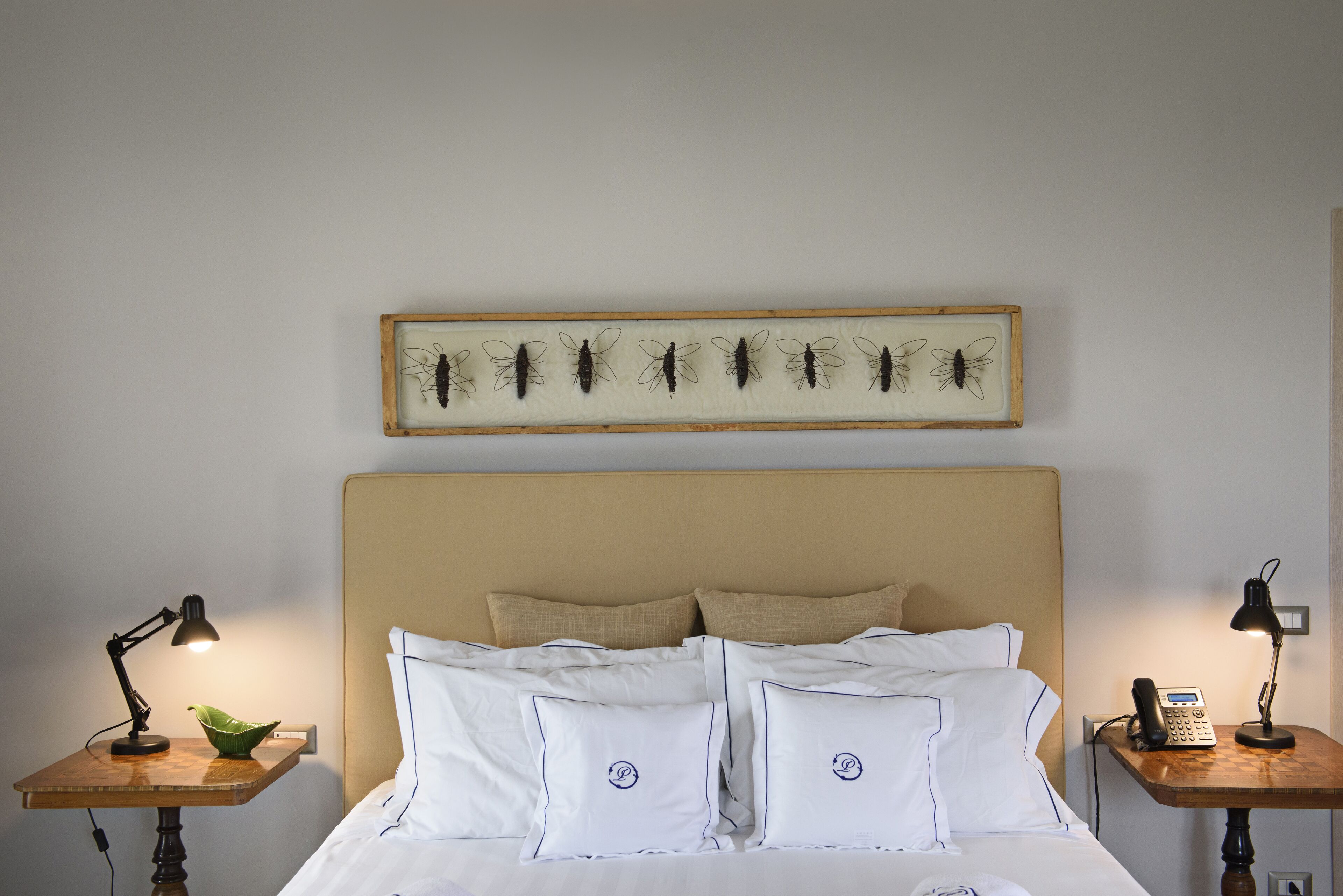 Un lit bien fait, avec du linge de lit blanc et des oreillers, deux tables de chevet avec des lampes, et une œuvre d’art encadrée représentant des insectes accrochée au mur.