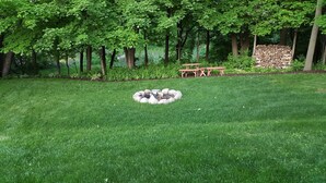 A well-maintained lawn with a stone fire pit in the center, surrounded by a wooden bench and a stack of firewood. - Natures Get-A-Way in Historical Excelsior (Excelsior)