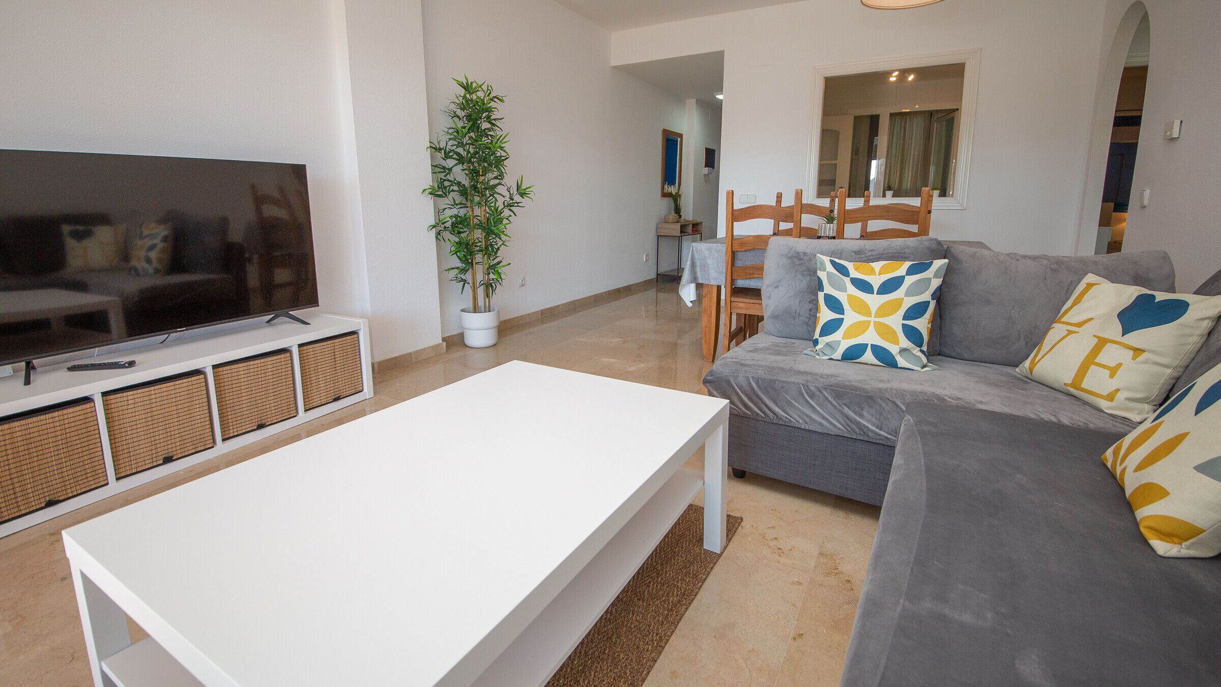 A modern living room with a flat-screen TV, a white coffee table, a grey sofa with patterned cushions, and a potted plant.