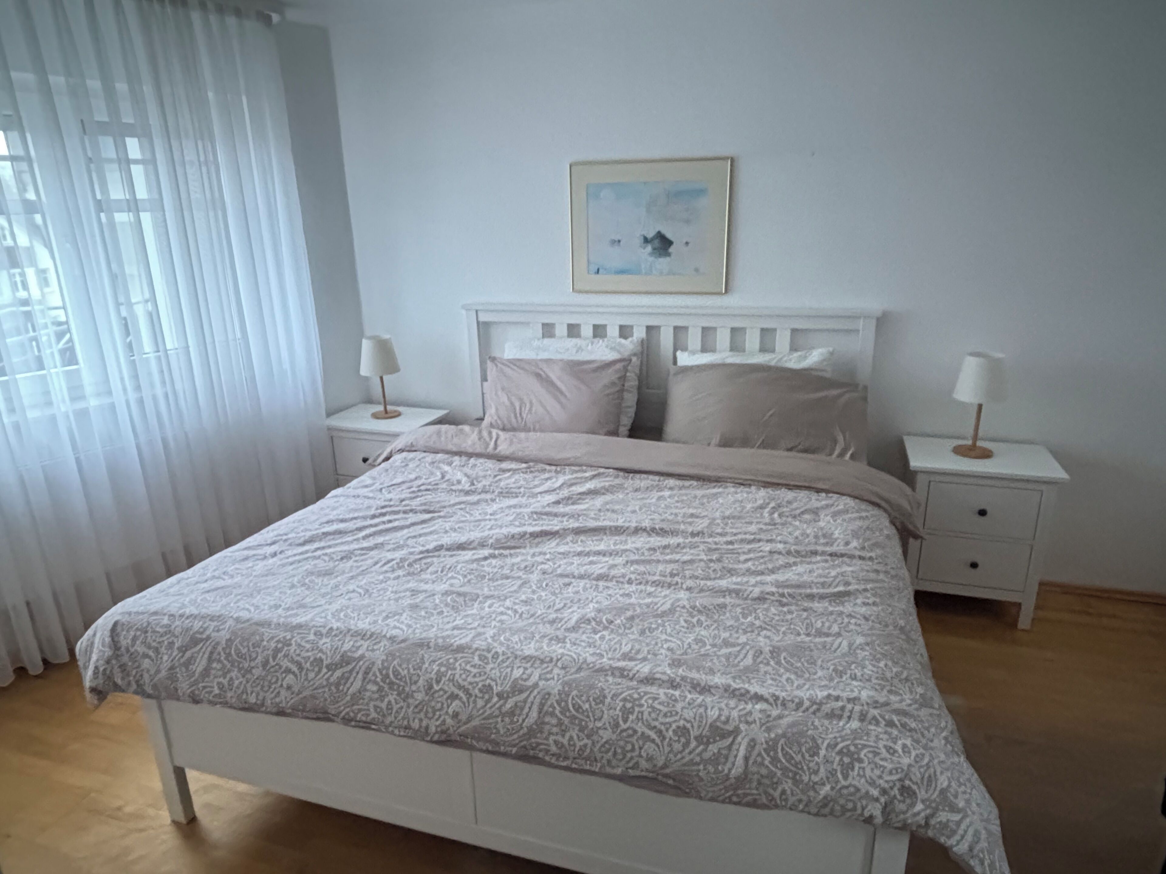 A bedroom with a bed, two bedside tables, a nightstand, a window with curtains, and a framed picture on the wall.