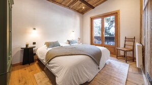 A bedroom with a wooden ceiling, a large bed with white bedding, a small bedside table, a chair, and a window with a view of trees. - Fully renovated La Datcha family chalet, ski room and playroom (Les Contamines-Montjoie)