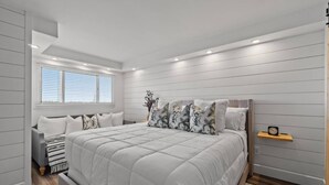 A bedroom with a large bed, a bedside table, a nightstand, a window with blinds, and a small shelf with a clock.