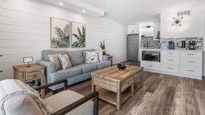 Una sala de estar moderna con un sofá gris, una mesa de centro de madera y una cocina con armarios blancos y un horno empotrado.