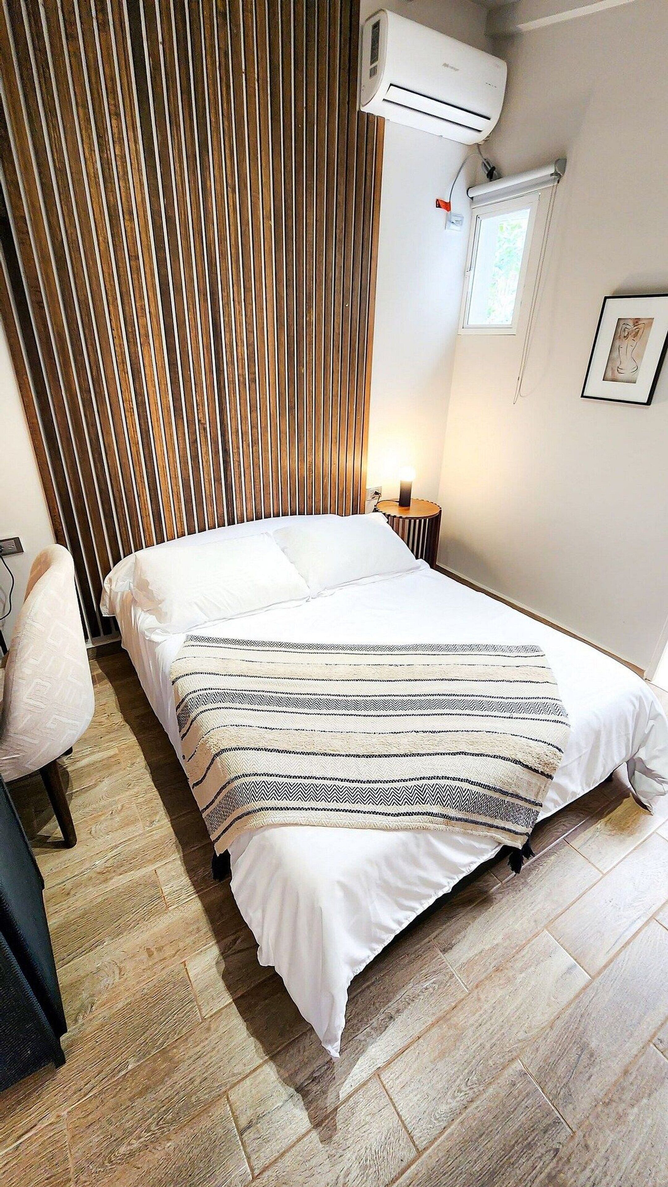 A hotel room with a bed, a bedside table with a lamp, a wooden slat wall, an air conditioning unit, and a framed picture on the wall.