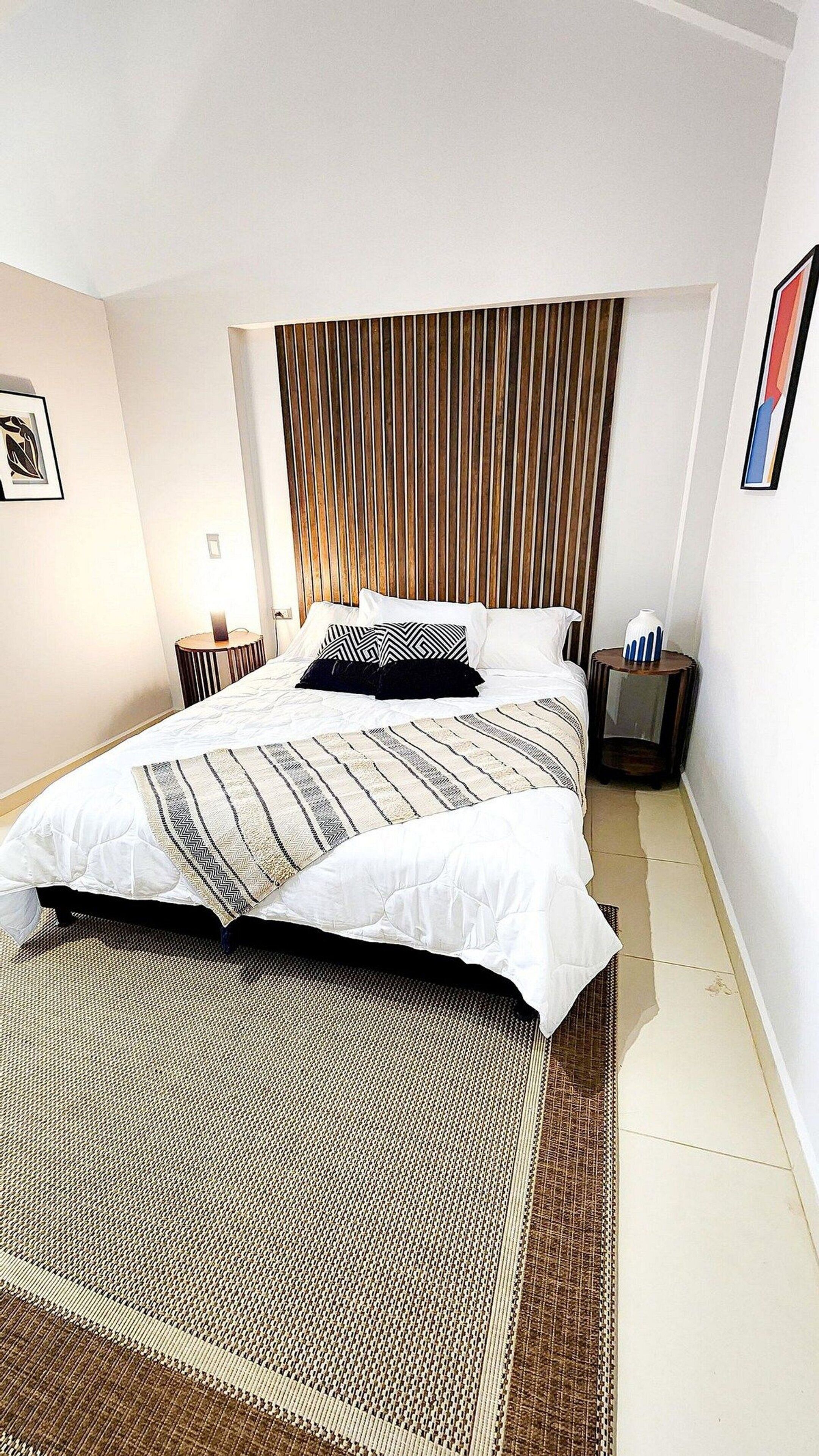 A modern bedroom with a large bed, a wooden slatted headboard, a patterned rug, and a small bedside table with a vase.