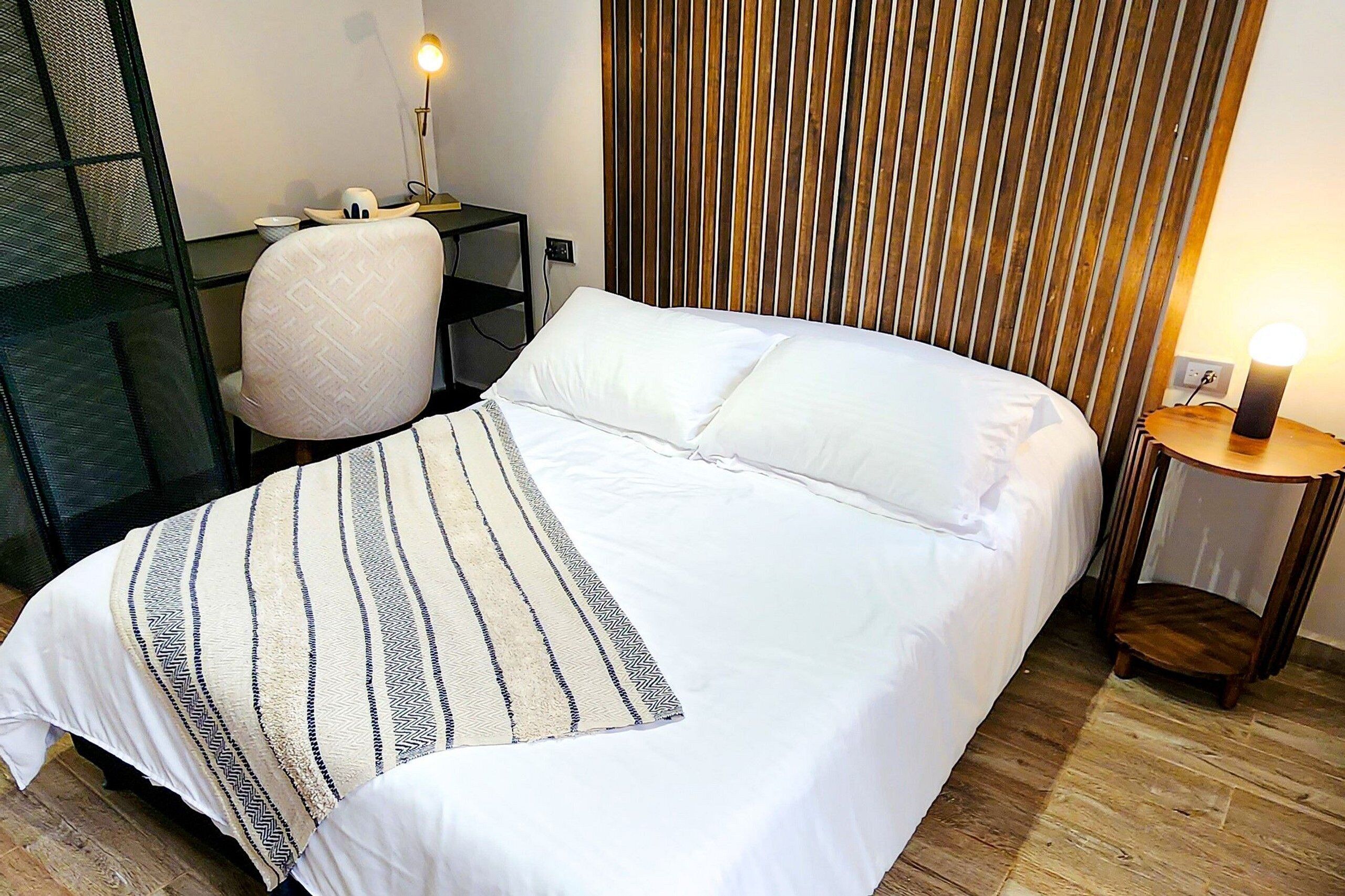 Een netjes opgemaakt bed met een gestreept dekbed, een houten lattenbovenstuk, een bureau met stoel en een nachtkastje met lamp.