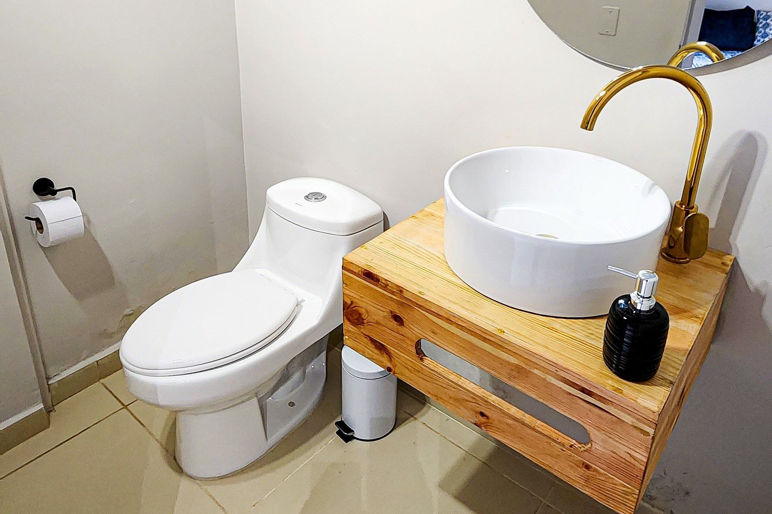 A bathroom with a white toilet, a wooden vanity with a white sink and gold faucet, and a white trash bin.