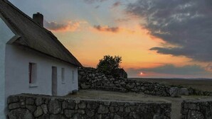 A white building with a thatched roof, a stone wall, and a view of the sunset. - Historical Thatched Cottage @ Multi-Award-Winning Cnoc Suain (Galway)