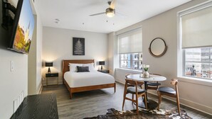 A hotel room with a bed, bedside tables, a desk, a chair, a round table, a ceiling fan, a window with a view, and a wall-mounted TV. - Bourbon Street Bliss (New Orleans)