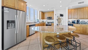 A modern kitchen with stainless steel appliances, wooden cabinets, a granite countertop island with stools, and large windows. - Stunning Lagoon Front Views | Pool | Fishing Pier & Boat Slips (Gulf Shores)