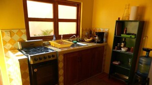 Cocina compacta con estufa a gas, horno y lavadero. Hay una ventana sobre el lavadero, un dispensador de agua y un estante verde con diversos utensilios de cocina.