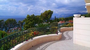 A curved pathway with a railing, surrounded by greenery and flowers, leading to a view of the sea and mountains.