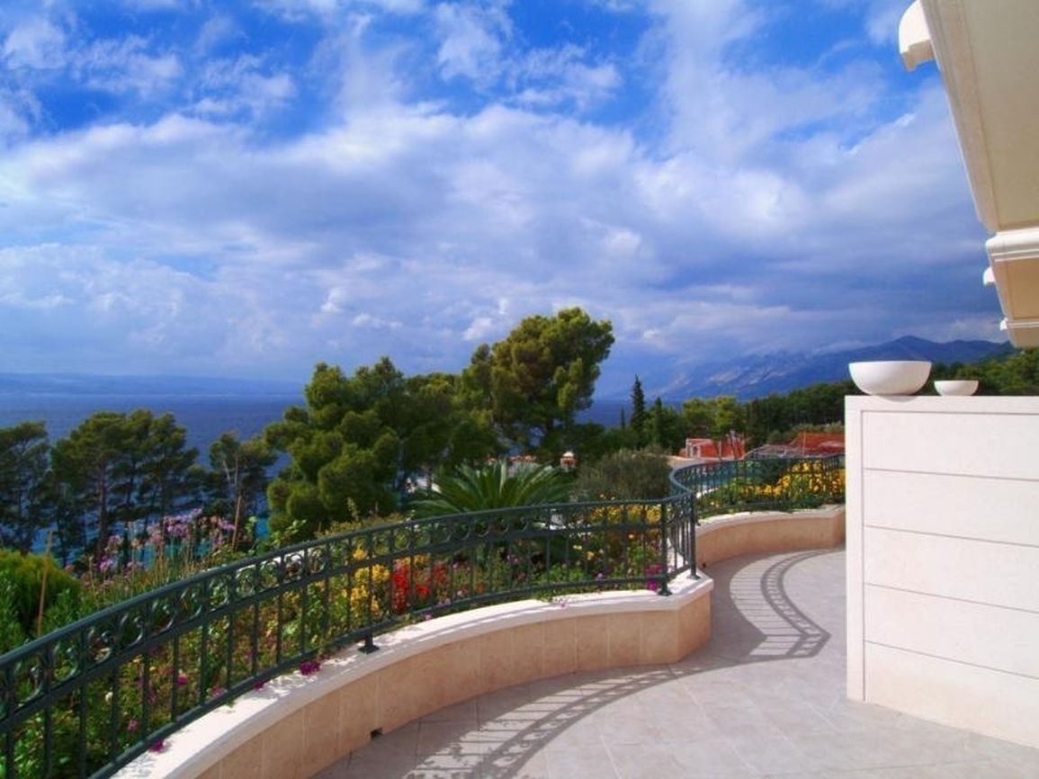 Un sendero curvo con barandilla, rodeado de follaje y flores, que conduce a una vista del mar y las montañas.