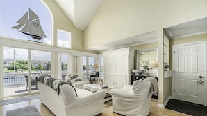 A spacious living room with a large window overlooking a balcony, white furniture, and a sailboat model. - HV Marina Condo with Boat Slip (Manistee)