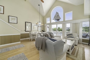 A spacious living room with a high ceiling, large windows, and a view of the outside. There is a dining area with a table and chairs, and a cozy seating area with a couch and armchairs.