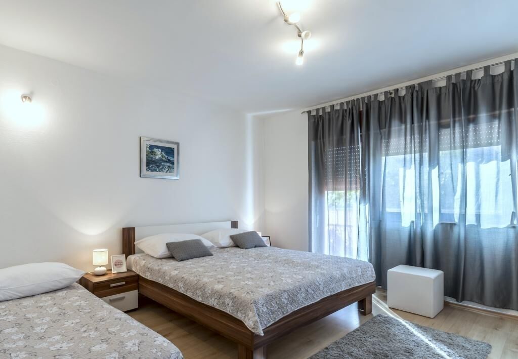 Quarto moderno com uma cama grande, mesas de cabeceira, uma mesinha de cabeceira com abajur, um criado-mudo e um quadro emoldurado na parede, além de uma janela com cortinas.