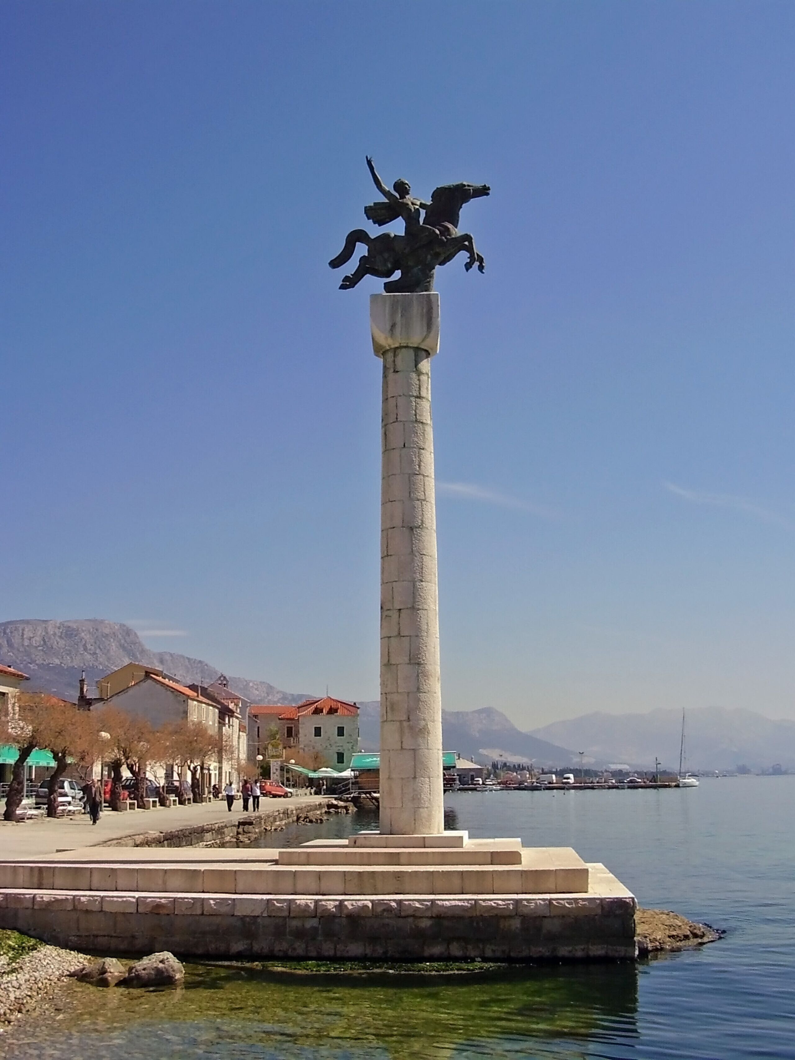 Un monumento alto con una estatua en la parte superior, situado cerca de un muelle con edificios y montañas al fondo.