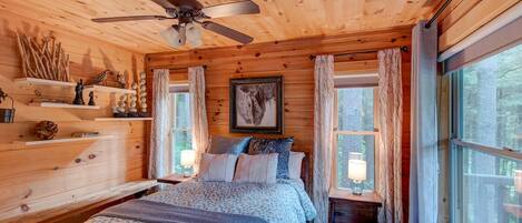 A cozy cabin bedroom with a wooden ceiling, a bed with patterned bedding, a ceiling fan, and a large window with curtains.