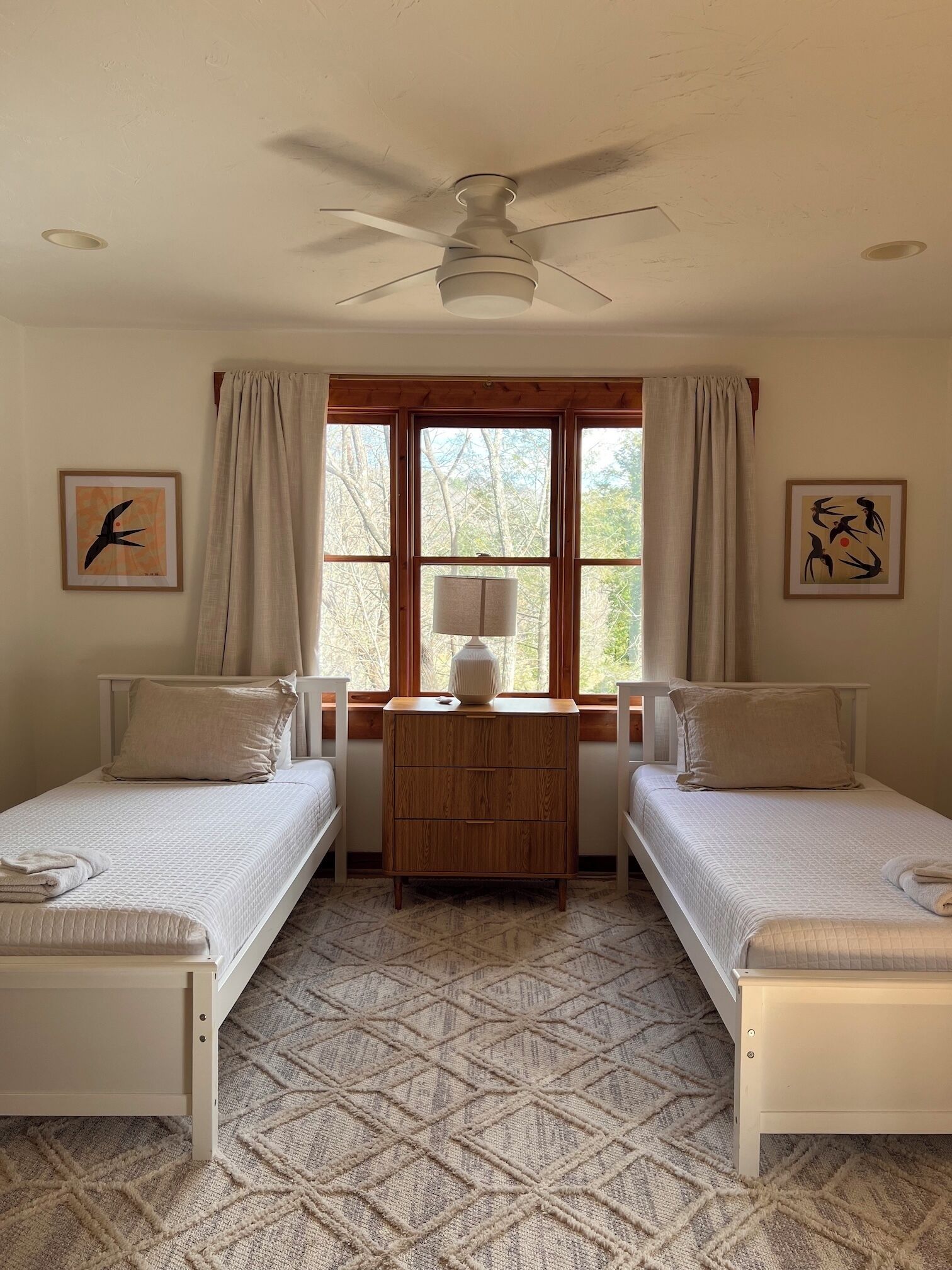 A room with two beds, a ceiling fan, a window with curtains, and artwork on the walls.